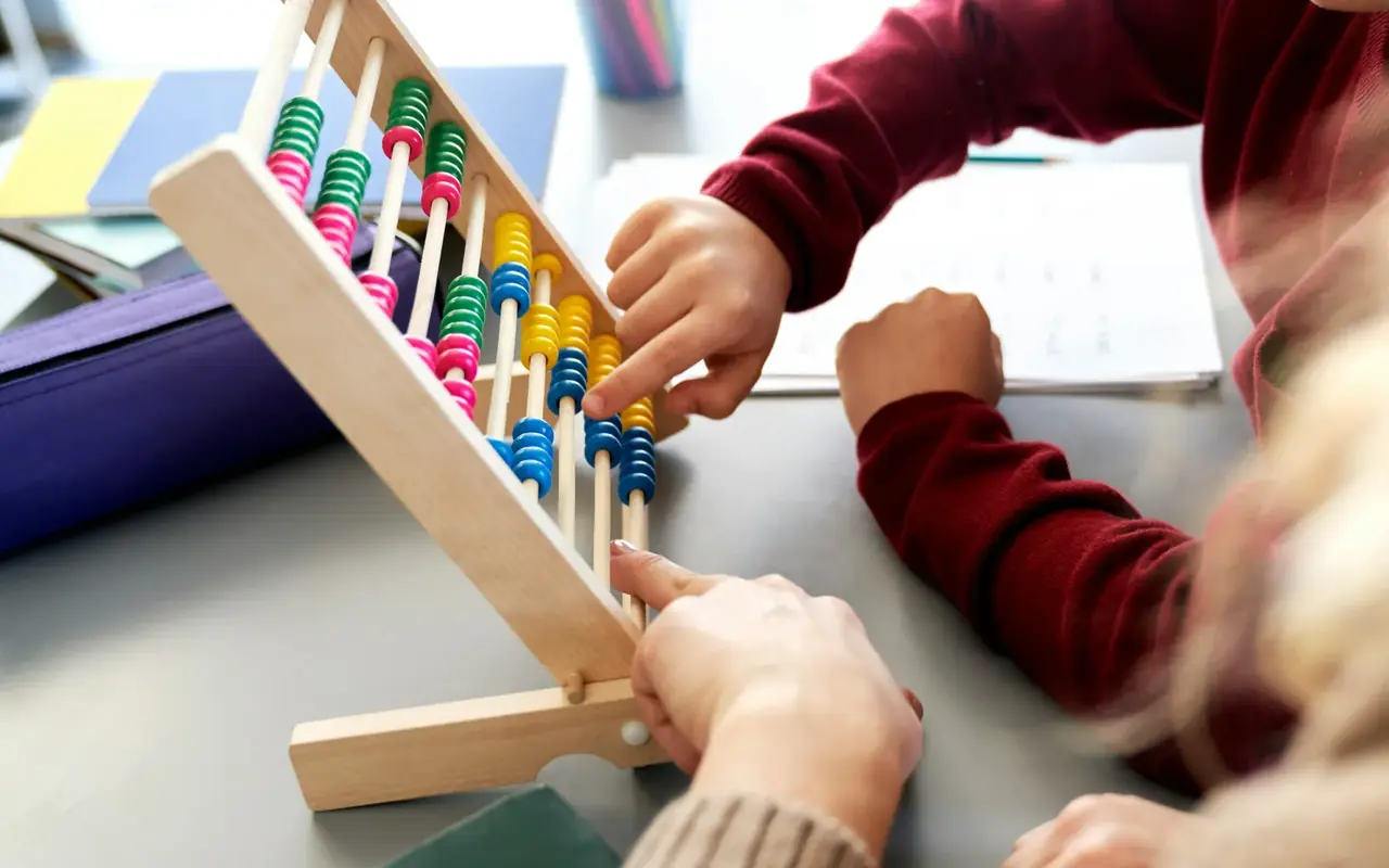 A teacher&rsquo;s and a student&rsquo;s hands working a wooden abacus together beside a paper worksheet