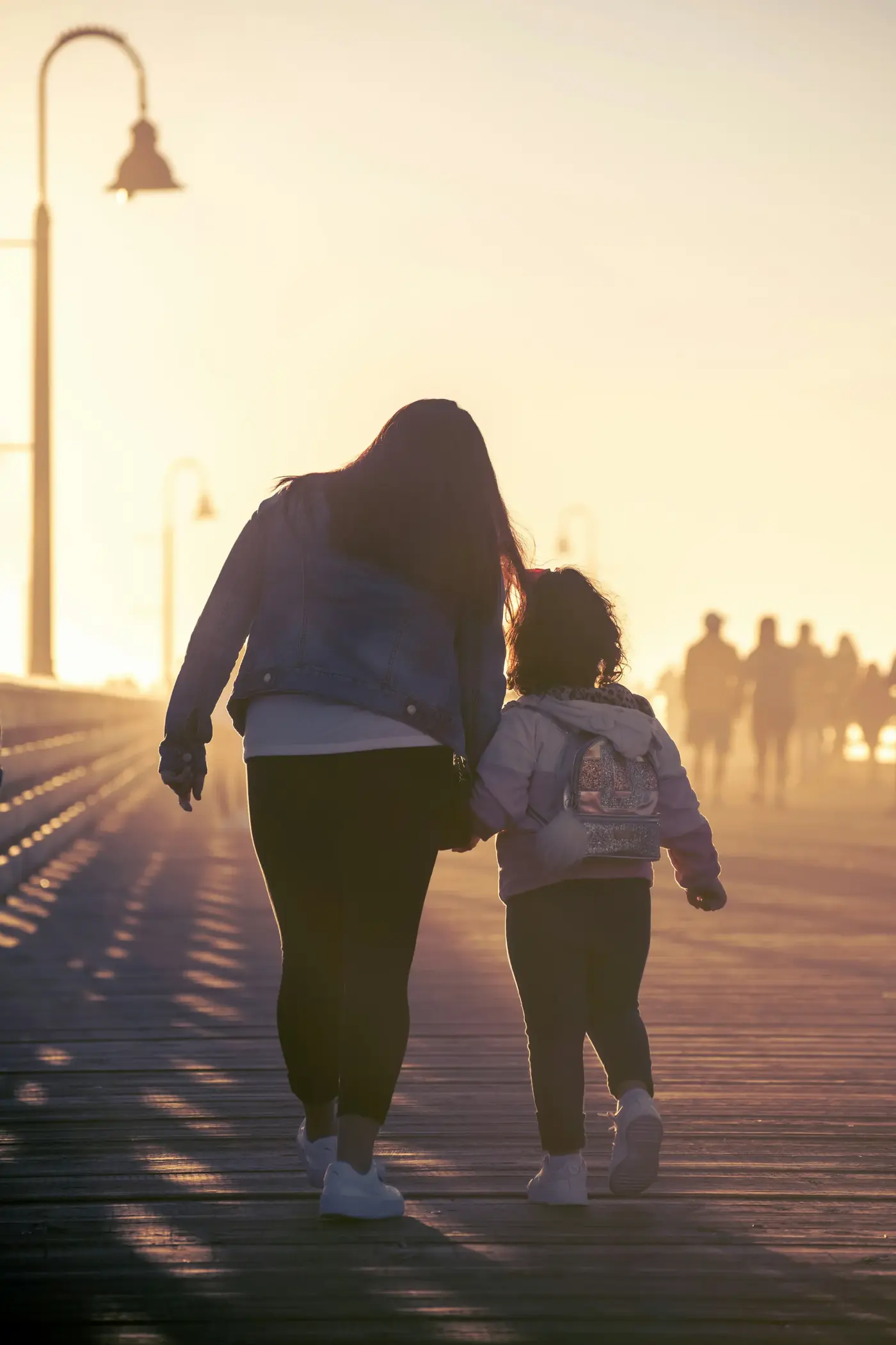 A parent and young child with a backpack walking hand in hand into golden morning light