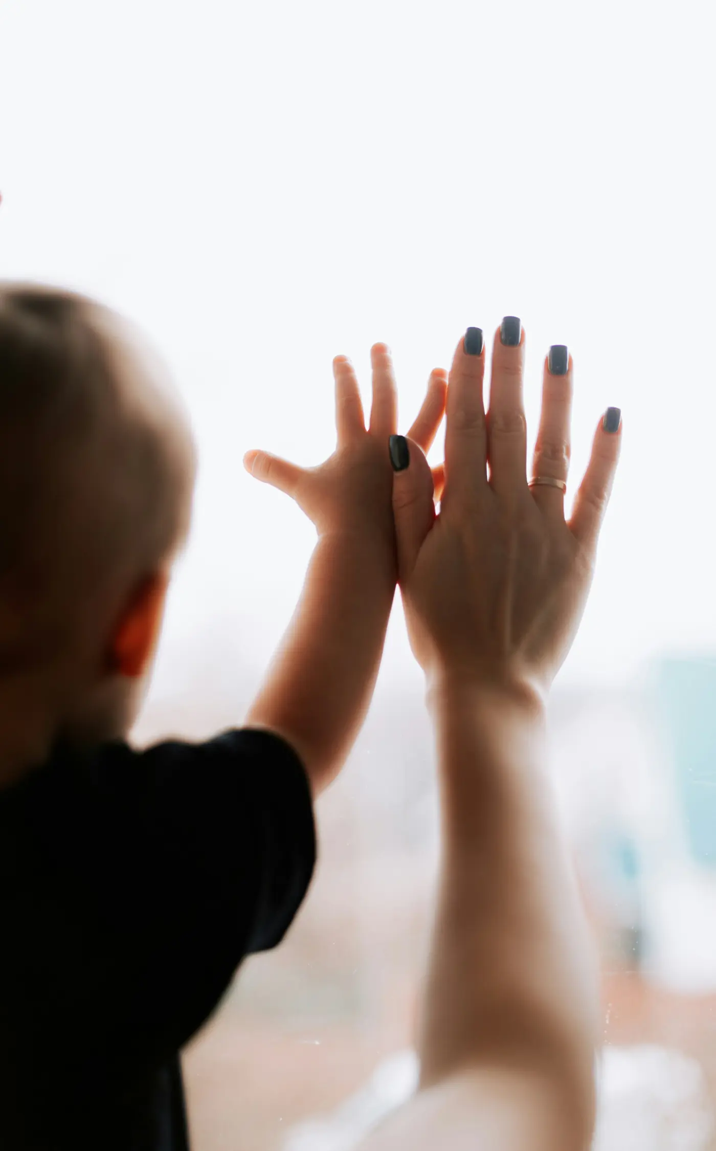 A young child&rsquo;s hand pressed against a parent&rsquo;s hand at a bright window
