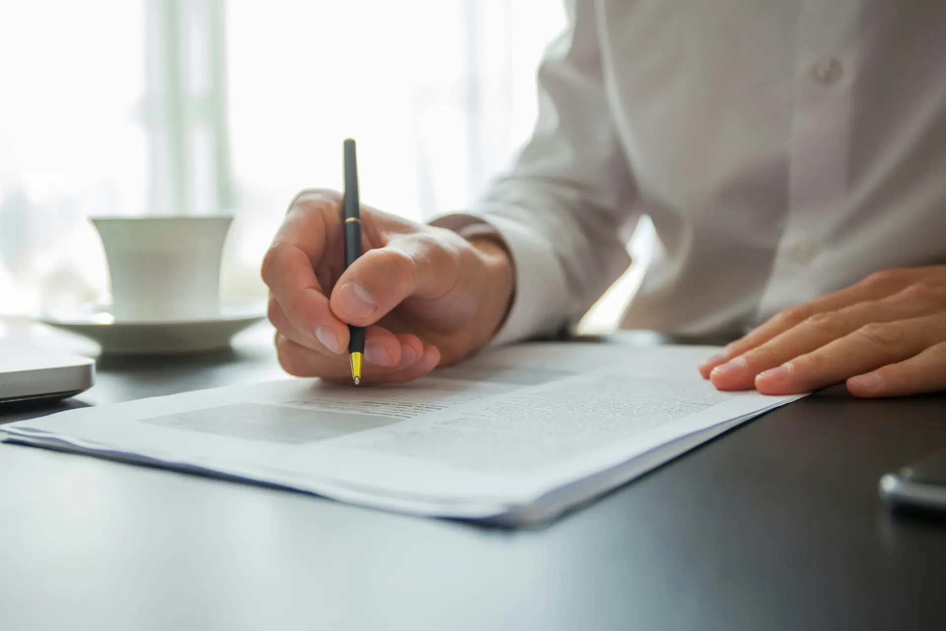A hand signing a printed document at a bright window, coffee alongside