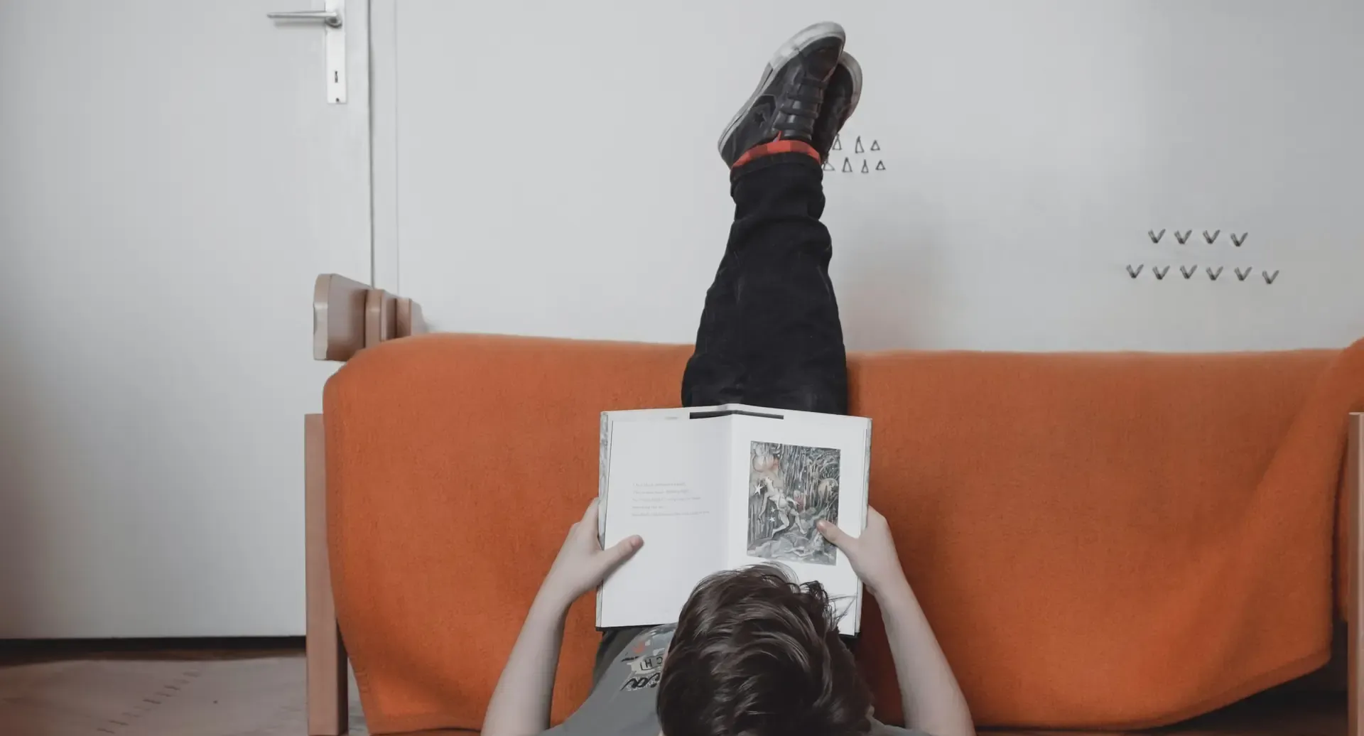 A boy lying on the floor reading, legs up on a couch, surrounded by open books